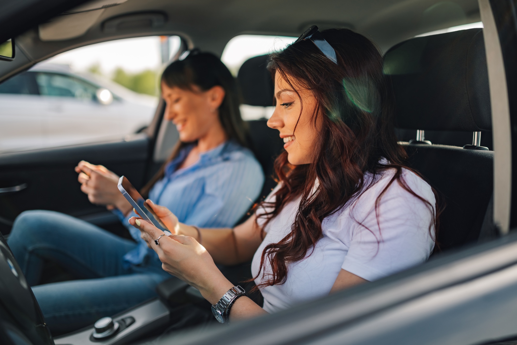Side view of happy traveler girls sitting in car with cellphones.